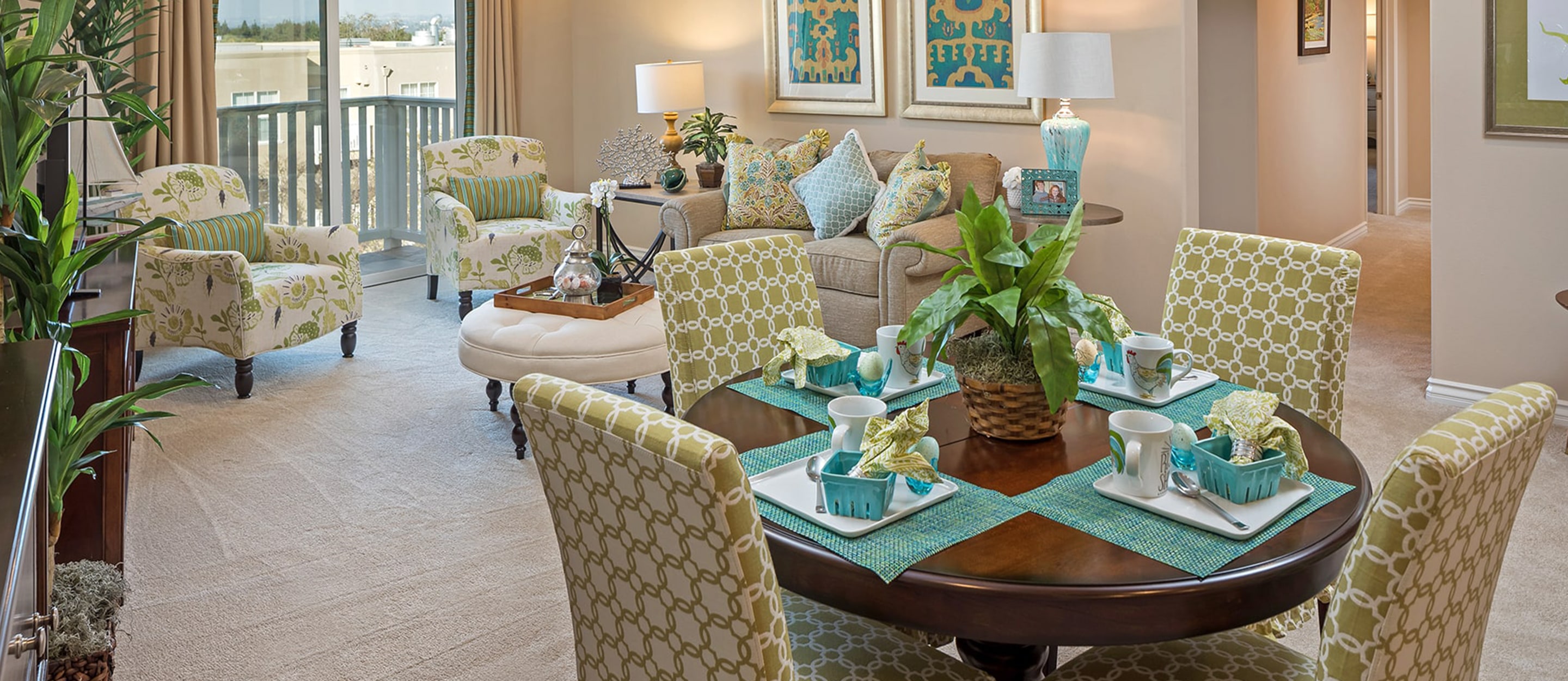 A bright, modern living and dining room with green and beige patterned chairs, a round dining table set for four, a cozy sofa, leafy plants, and large windows letting in natural light.