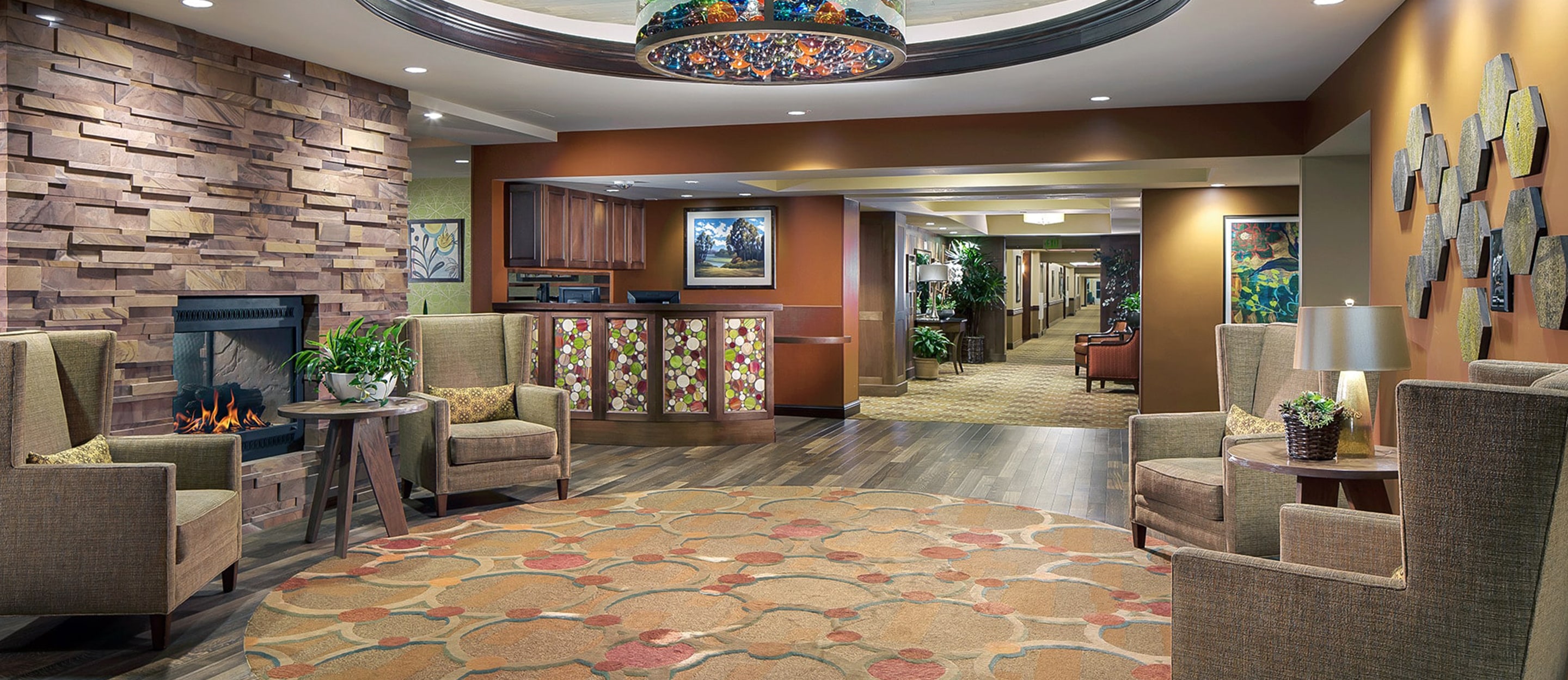 A cozy lobby with armchairs around a round rug, a lit fireplace, and a front desk. Warm lighting, earth-tone decor, wall art, and plants create a welcoming atmosphere. A hallway extends into the background.
