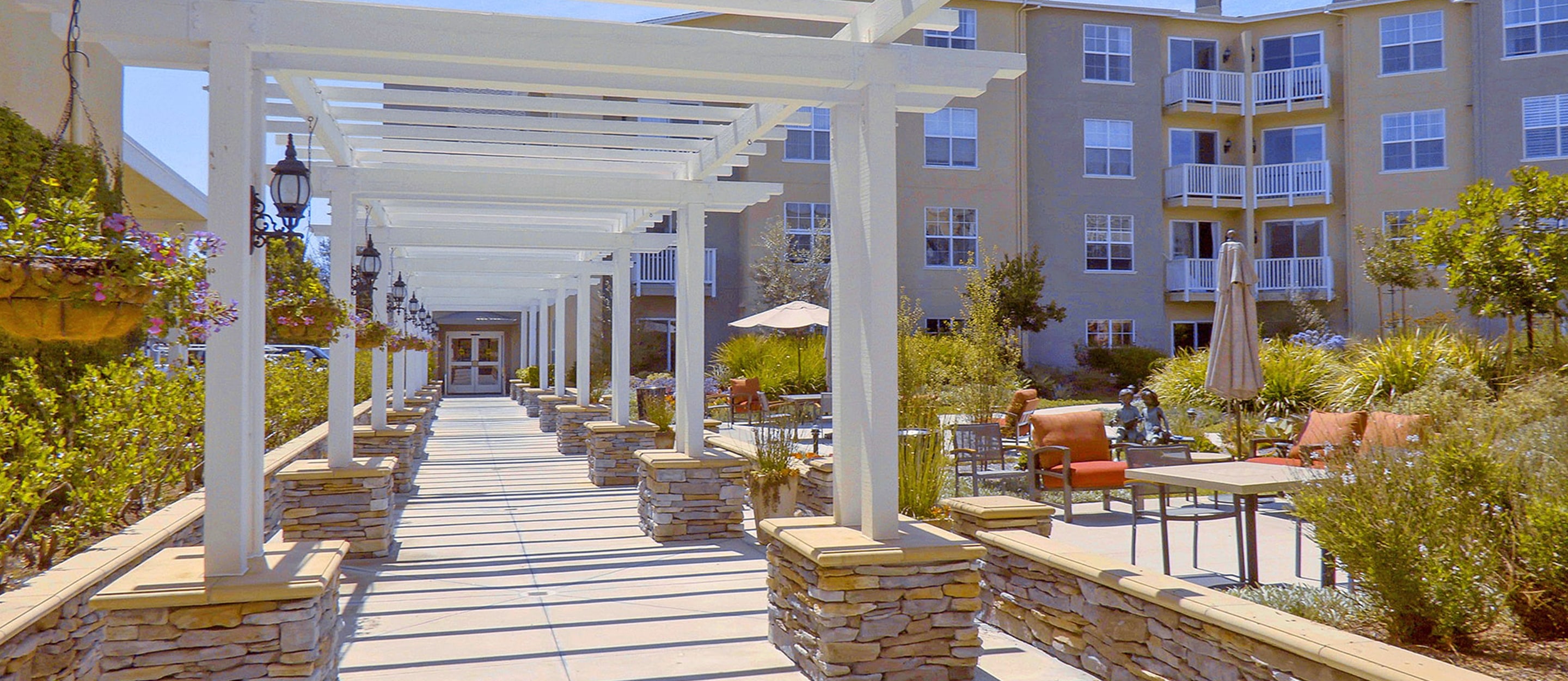 A wide walkway with a white pergola leads to an apartment building. The area is landscaped with greenery and patio seating, creating a welcoming outdoor space.