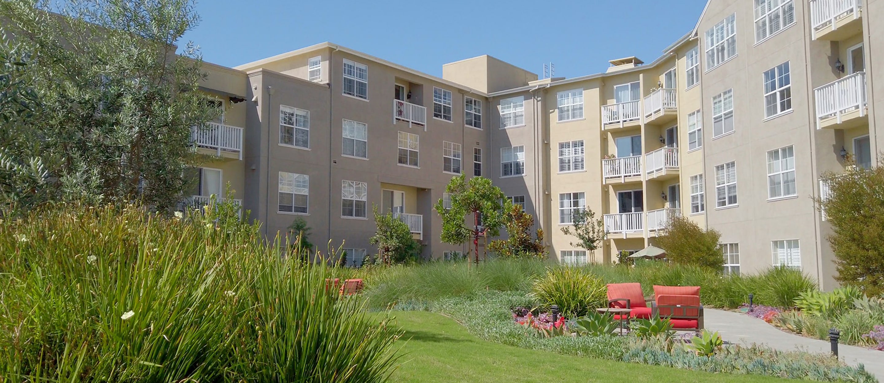 A modern three-story apartment building with beige exterior walls, surrounded by green lawns, plants, and red outdoor seating under a clear blue sky.