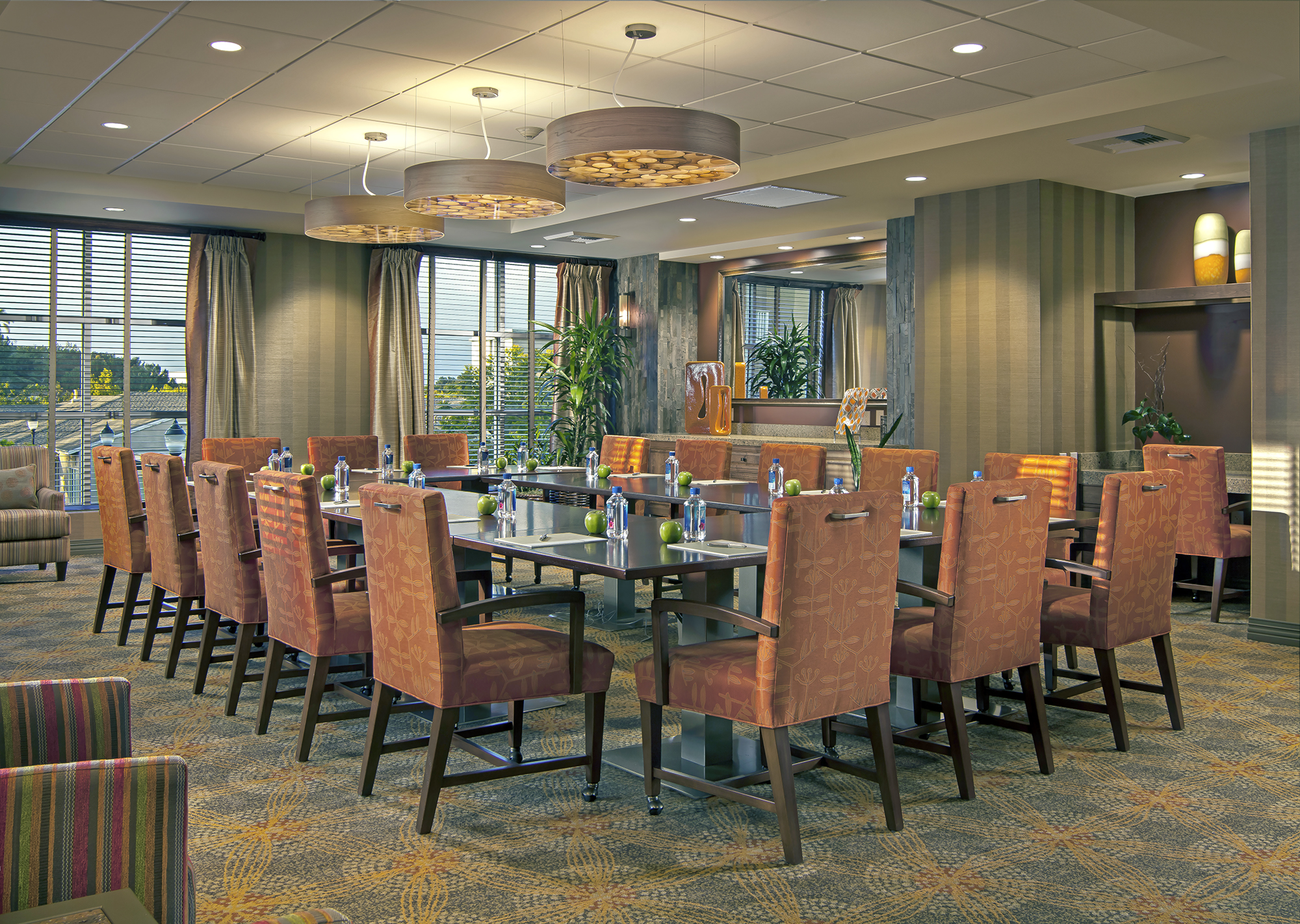 A well-lit conference room with large windows, arranged tables in a U-shape, orange upholstered chairs, bottled water, notepads, contemporary light fixtures, and green plants in the background.