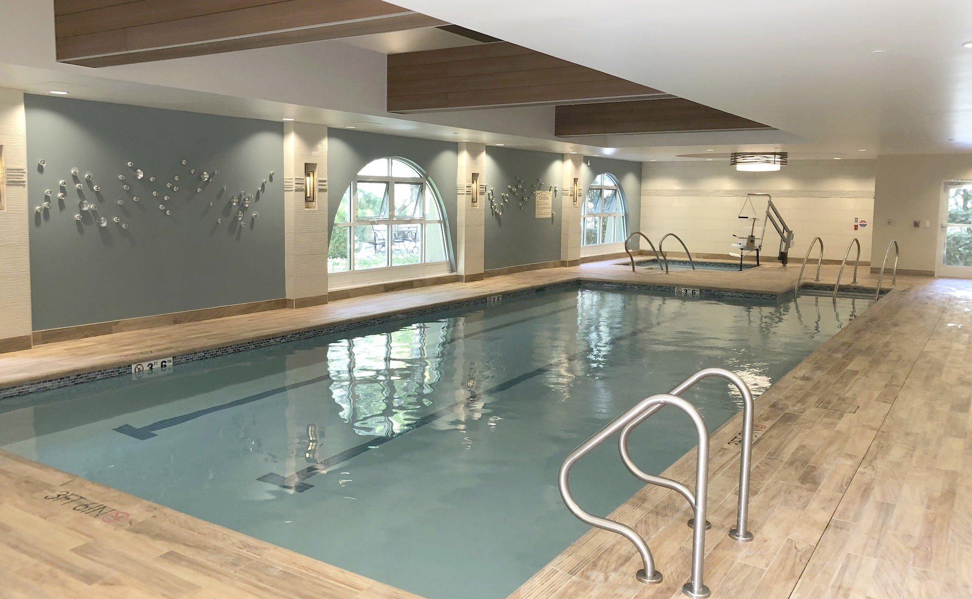 Indoor swimming pool with wood-like tile flooring, metal handrails, decorative wall art, large arched windows, and soft overhead lighting in a modern, clean, and inviting space.