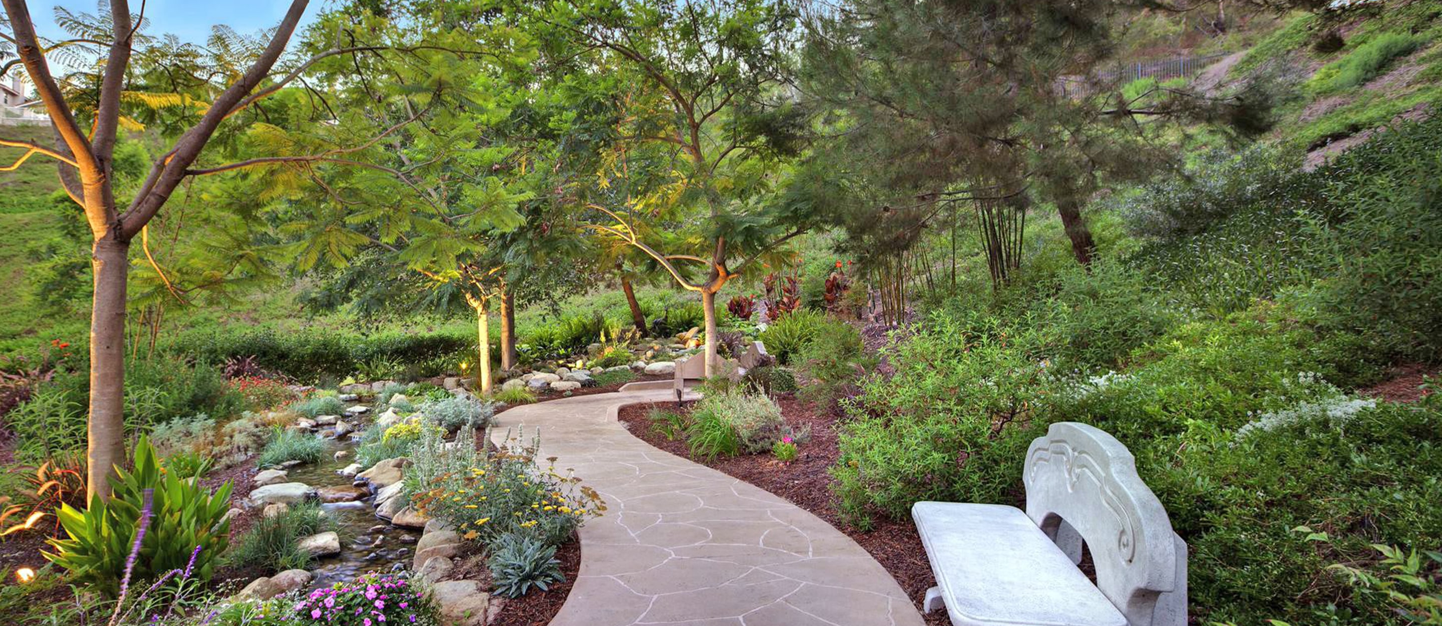 Curving stone path winds through a lush garden with trees, shrubs, flowers, a small creek with rocks, and a decorative stone bench on the right, creating a serene, peaceful outdoor setting.