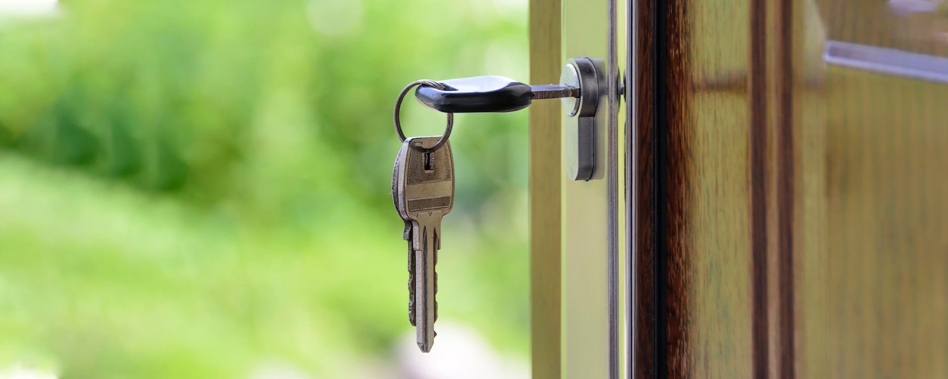 A close-up of a door slightly open with a key inserted in the lock. Two additional keys are attached to the keyring. The background is blurred greenery, suggesting an outdoor garden or yard.