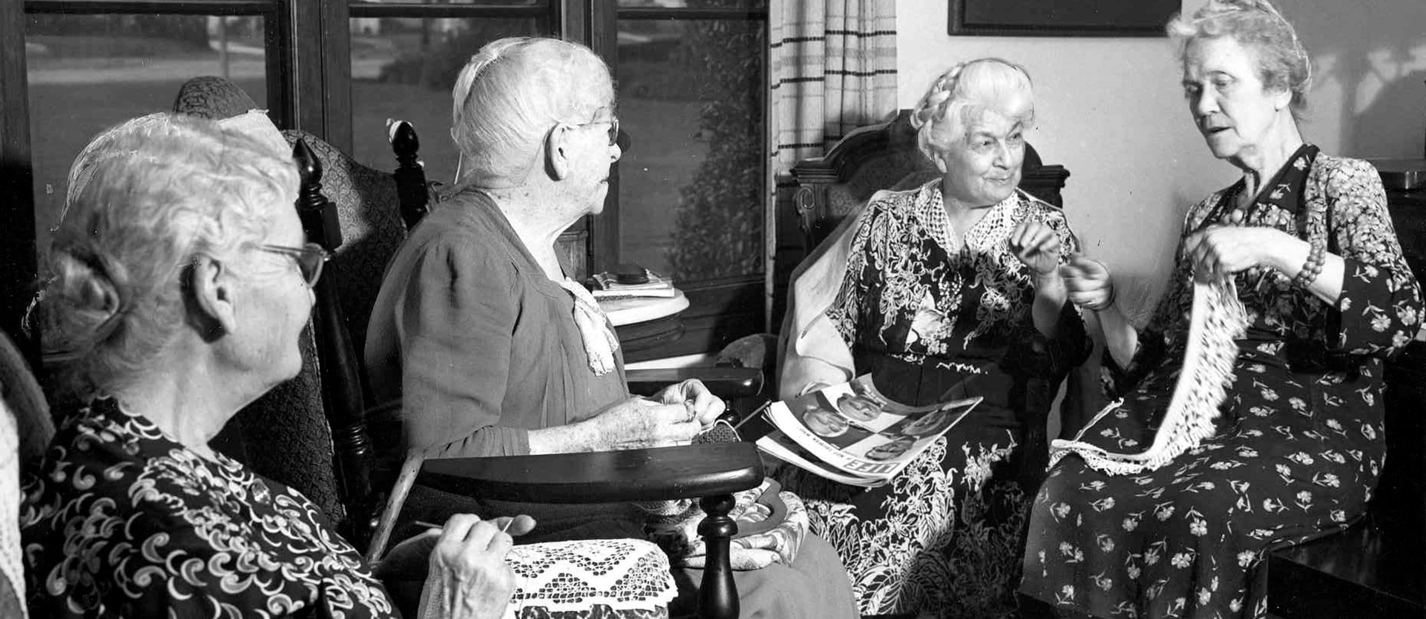 Four elderly women sit together in a living room, talking and knitting. One woman holds a magazine, while another is working with yarn. They are dressed in patterned dresses and appear to be enjoying each other’s company.