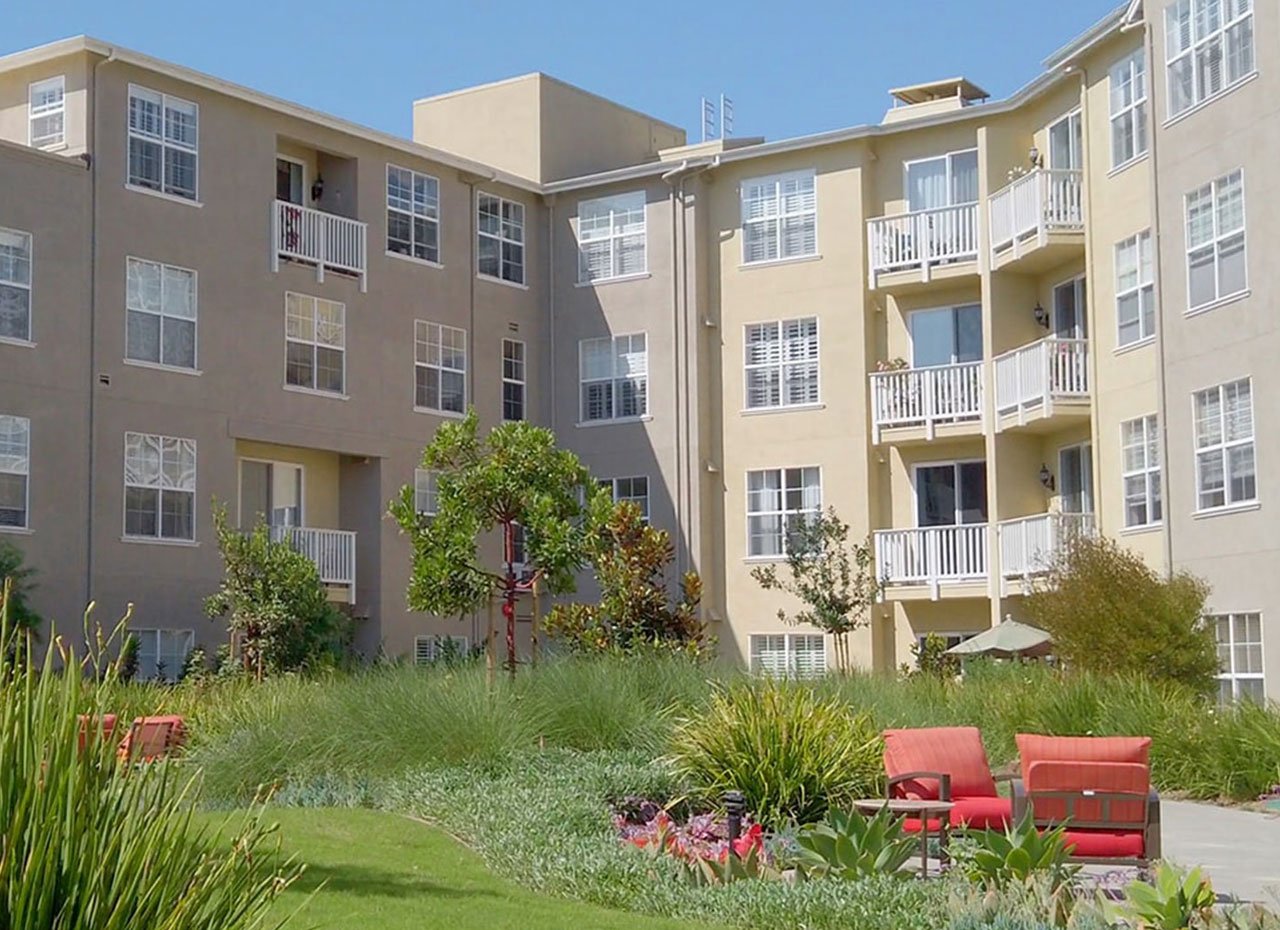 Three-story beige apartment buildings with white trim surround a landscaped courtyard featuring green grass, shrubs, flowers, small trees, and red outdoor seating under a clear blue sky.