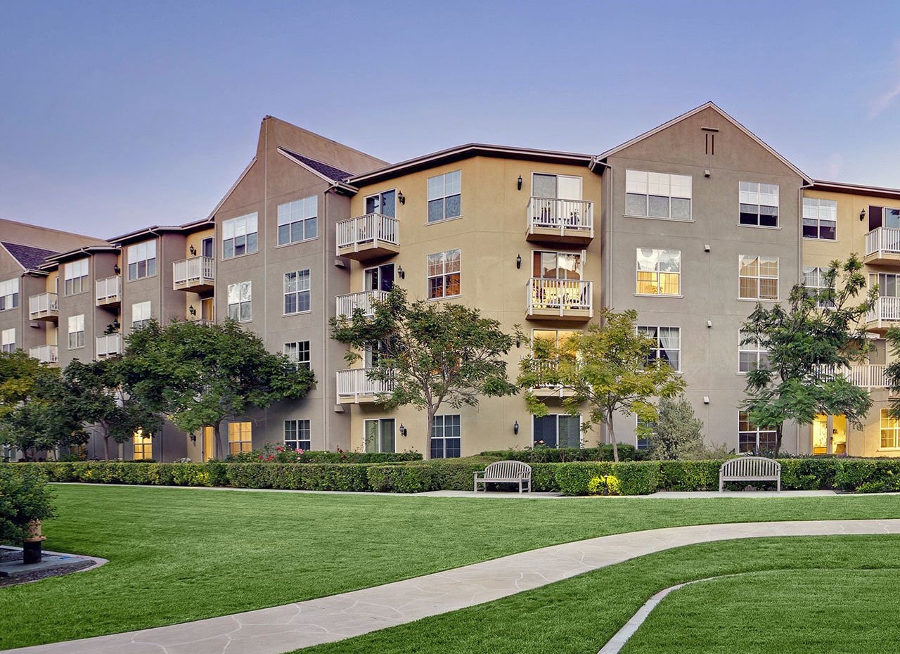 Four-story apartment building with beige exterior, balconies, and large windows, surrounded by green lawns, trees, benches, and a winding sidewalk under a clear sky.