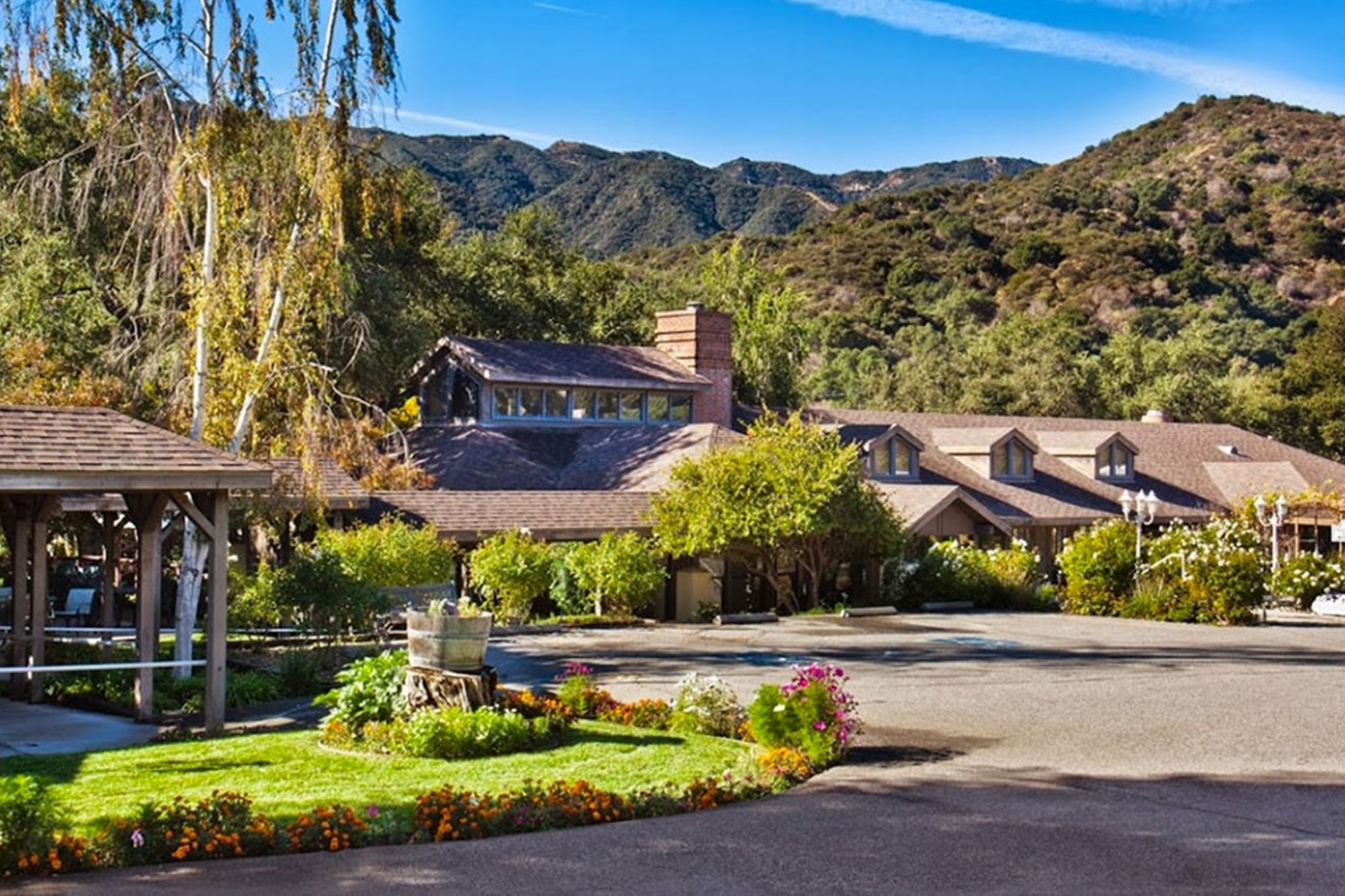 A rustic lodge surrounded by lush greenery and colorful flowers, set against a backdrop of tree-covered hills and a clear blue sky on a sunny day.