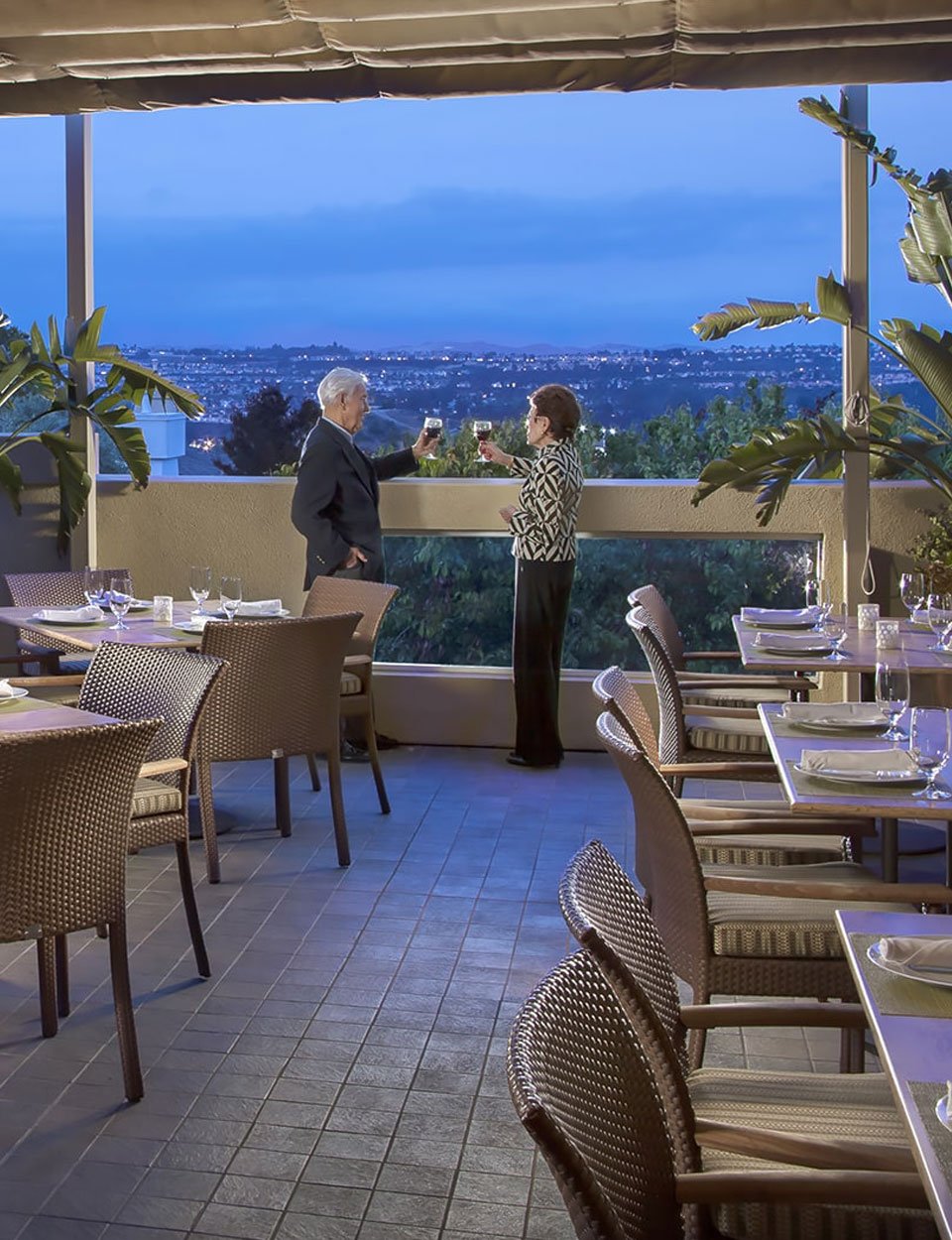 An older man and woman stand on a restaurant terrace at sunset, clinking glasses. Empty tables with place settings surround them, and a cityscape is visible in the background. Large plants decorate the area.