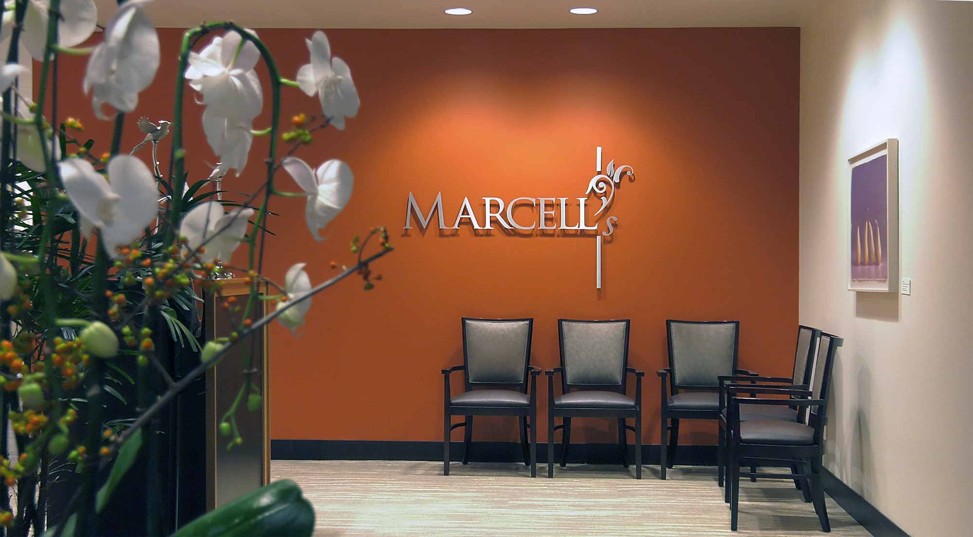 A waiting area with four chairs against an orange wall featuring the word MARCELLS in large letters. White orchids are in the foreground, and a framed picture hangs on the right wall.