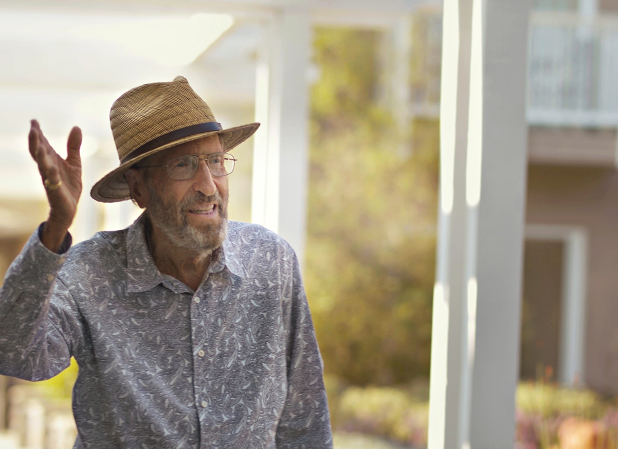 An elderly man wearing a straw hat, glasses, and a patterned shirt gestures with one hand while standing outdoors near a building, with soft sunlight in the background.