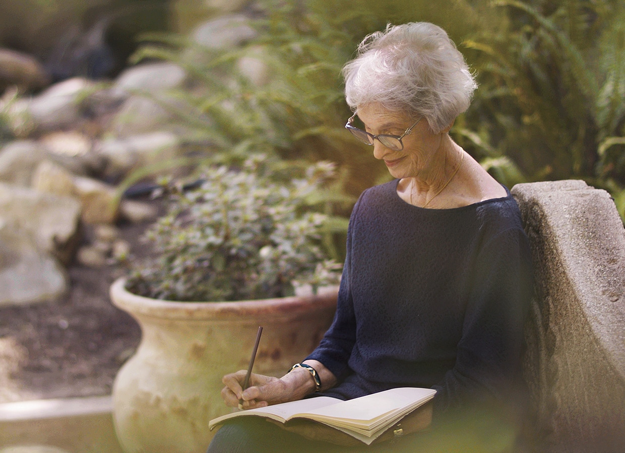 An older woman with short gray hair and glasses sits outdoors on a stone bench, smiling as she writes in an open notebook. There are plants and a large pot in the background, creating a peaceful garden setting.