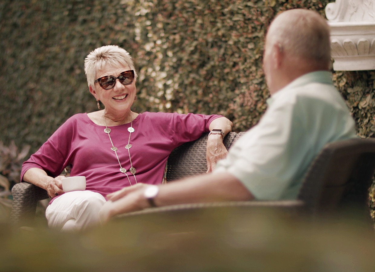 An older woman wearing sunglasses and a purple top smiles while sitting outdoors on a wicker chair, talking to an older man. She holds a cup, and greenery fills the background.
