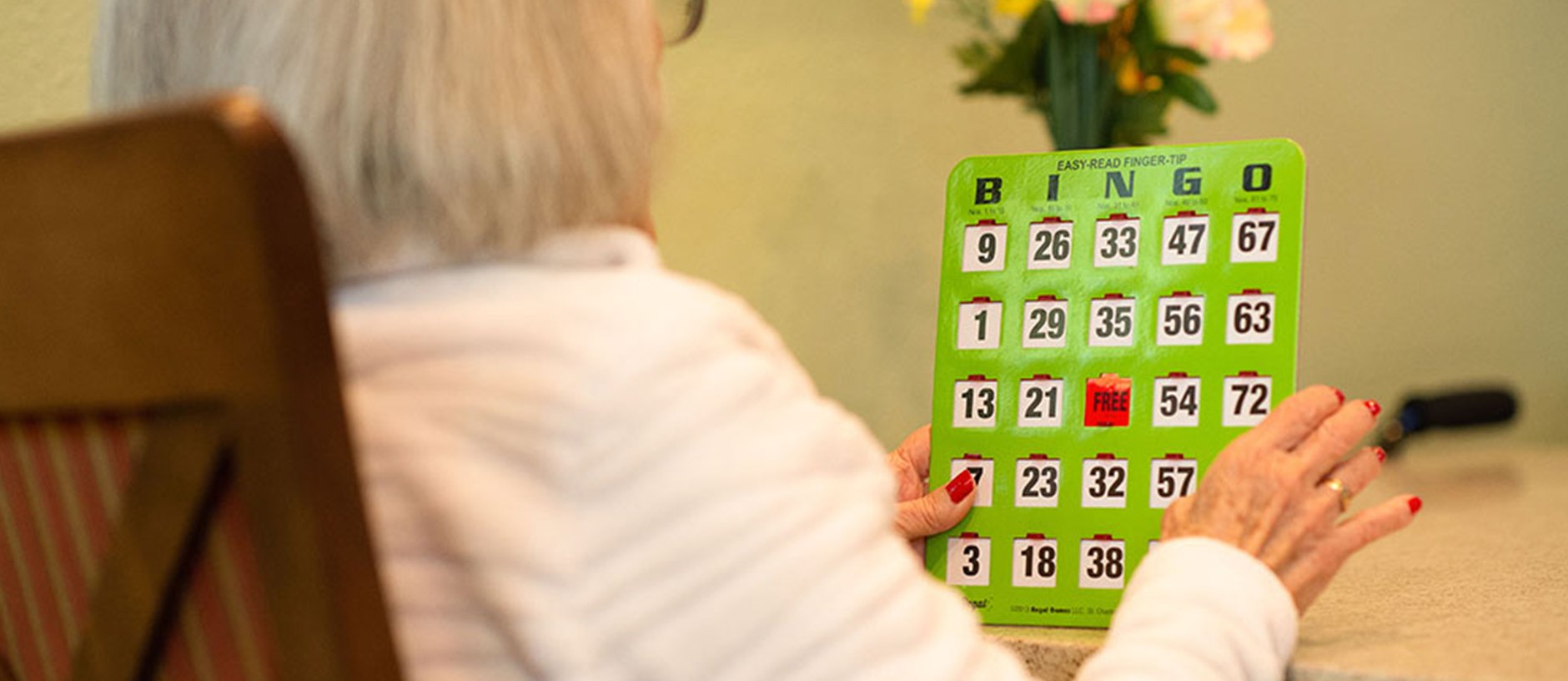 An older adult with light hair and a white top holds a green bingo card while sitting at a table. The persons face is not visible. A vase with yellow flowers is in the background.