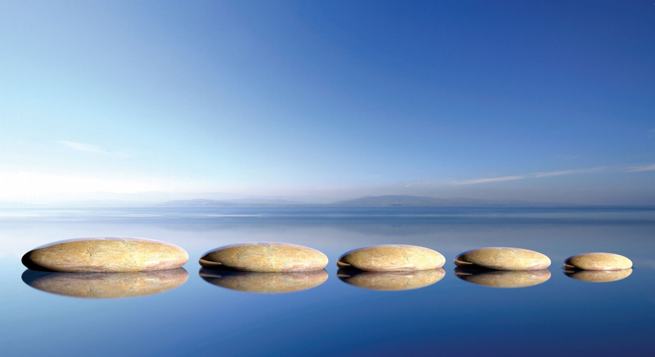 Six smooth, oval stones are arranged in a line on a calm, reflective water surface under a clear blue sky, creating a serene and peaceful atmosphere. Mountains faintly appear in the distant background.