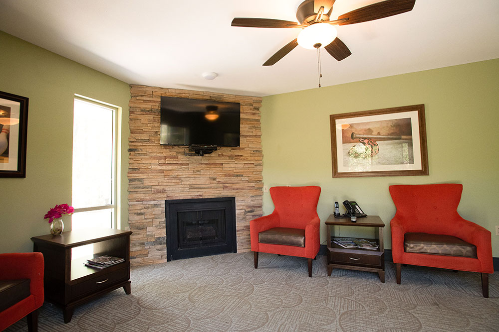 A cozy living room with two bright orange chairs, a fireplace with a stone surround, a wall-mounted TV, a ceiling fan, and side tables with flowers, phones, and magazines. Soft natural light enters from a window.