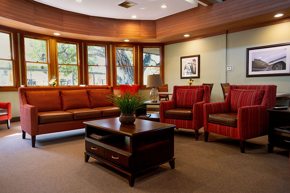 A cozy living room with a brown sofa, two red armchairs, a wooden coffee table with a plant, large windows letting in natural light, and framed pictures on the walls.