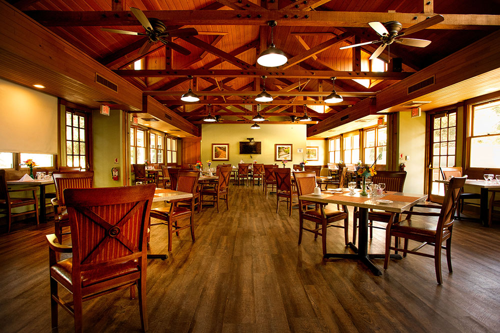 A spacious, warmly lit restaurant with wooden floors, exposed beams, ceiling fans, and large windows. Tables are set for dining, each with chairs and simple table settings, creating a cozy and inviting atmosphere.