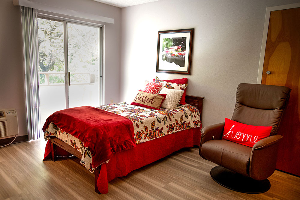 A cozy bedroom with a single bed decorated in red and floral bedding, throw pillows reading “blessed” and “home,” a brown recliner chair, wooden flooring, a framed picture, and a sliding glass door letting in light.