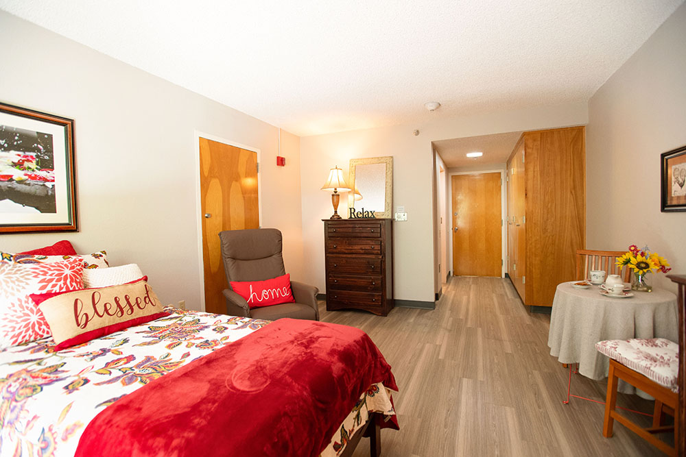 A cozy, well-lit bedroom with a bed covered in red and floral bedding, pillows labeled blessed and home, a recliner, a dresser with a lamp, a small round table with flowers, and wooden doors and floors.