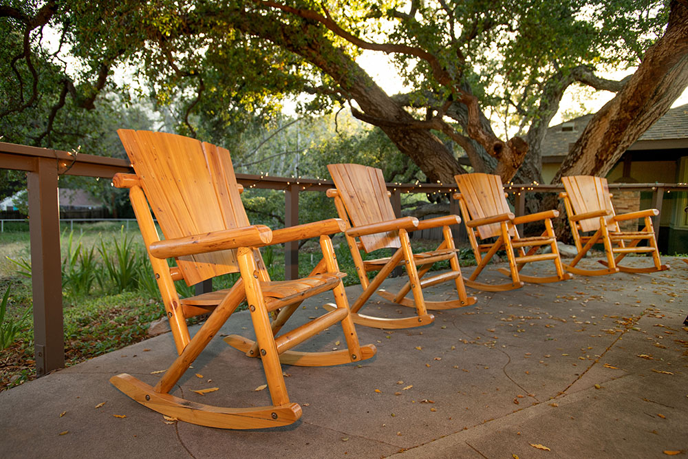 Four wooden rocking chairs are lined up on a patio beneath large, leafy trees. String lights hang along the railing, and sunlight filters through the branches, creating a peaceful, inviting outdoor setting.