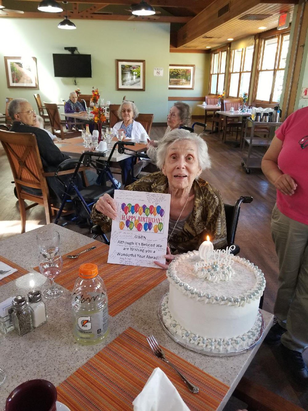 An elderly woman in a wheelchair smiles and holds a colorful birthday sign at a decorated table with a birthday cake and lit candle. Other people sit nearby in a cozy, sunlit room.