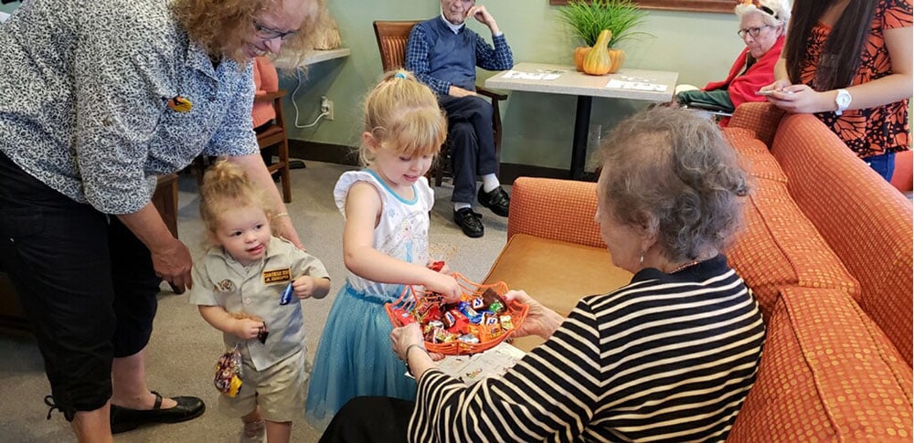 Two young children, one in a light blue dress and one in a tan outfit, offer candy to an elderly woman seated on an orange couch in a living room. Other adults watch and smile in the background.