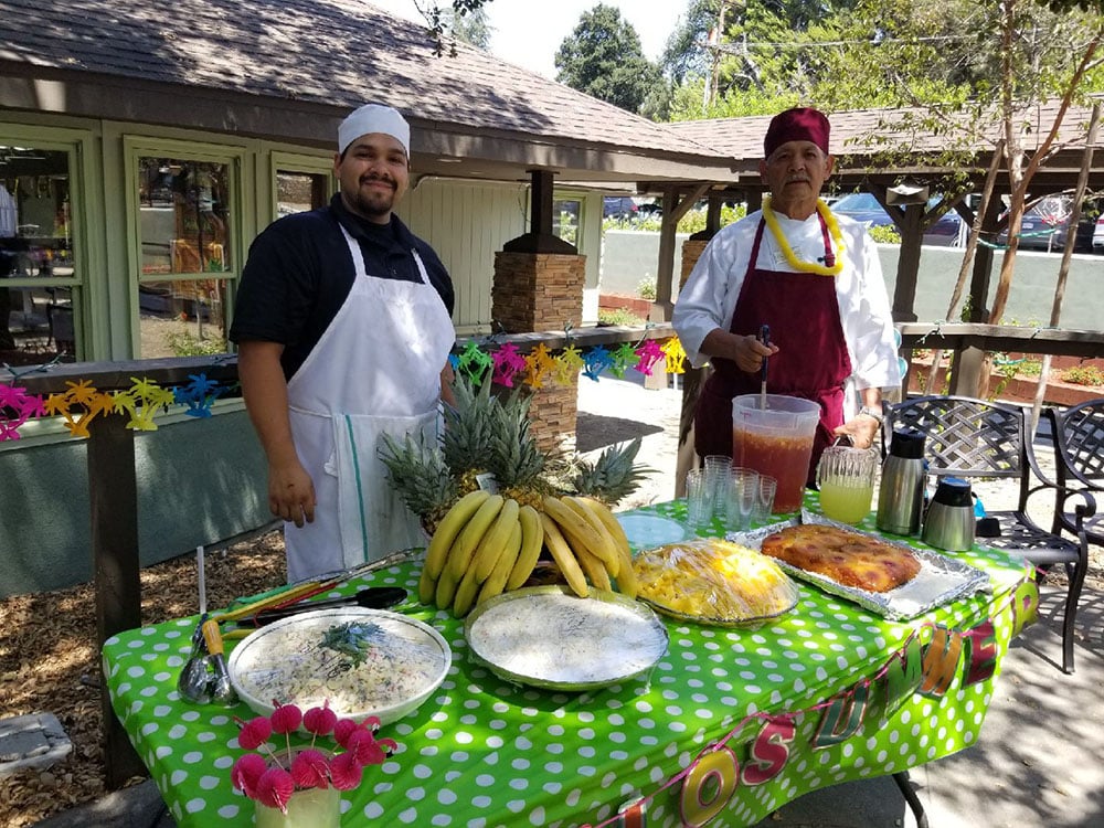 Two men in chef uniforms stand behind a table covered with a green polka dot cloth, offering food, fruit, and drinks outdoors. Colorful decorations and trees are visible in the background.
