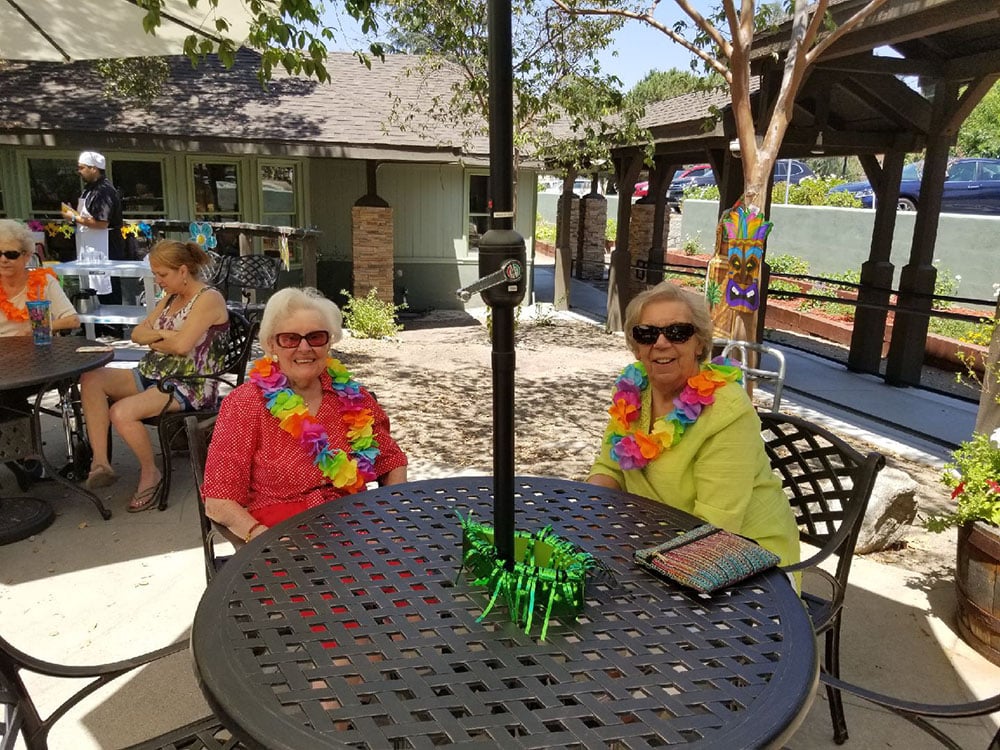 Two women wearing colorful leis and sunglasses sit at a round outdoor table, smiling. Other people are in the background, sitting at tables near a building, on a sunny day. A green decoration is on the table.