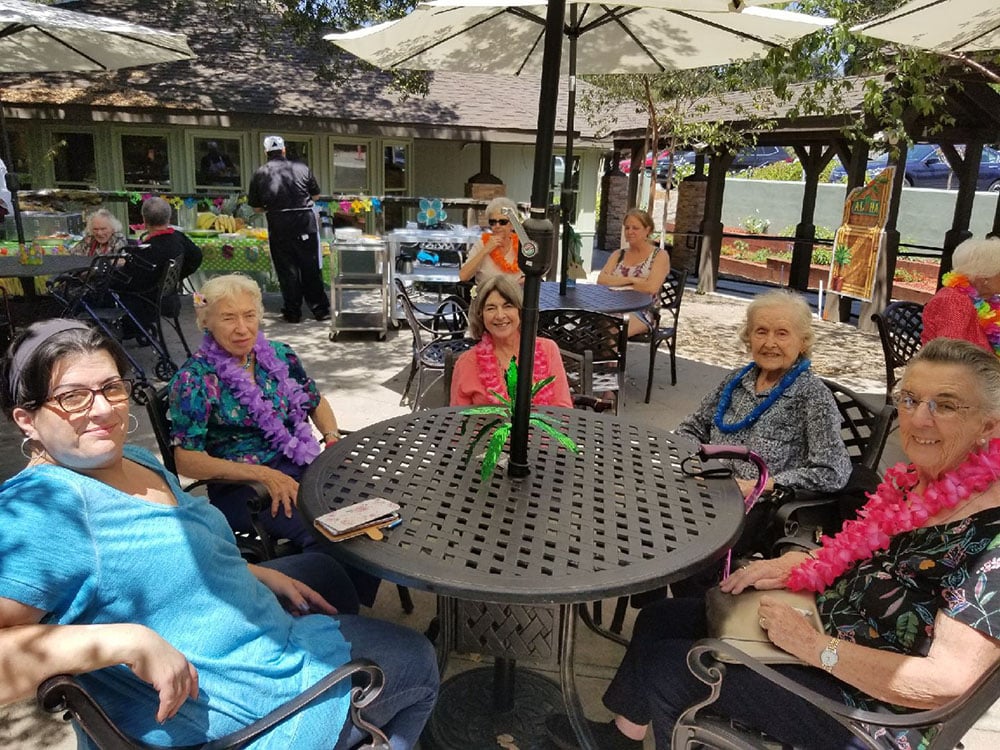 A group of older women wearing colorful leis sit around a patio table outdoors under an umbrella, smiling. Other people and decorations are visible in the background at a festive gathering.