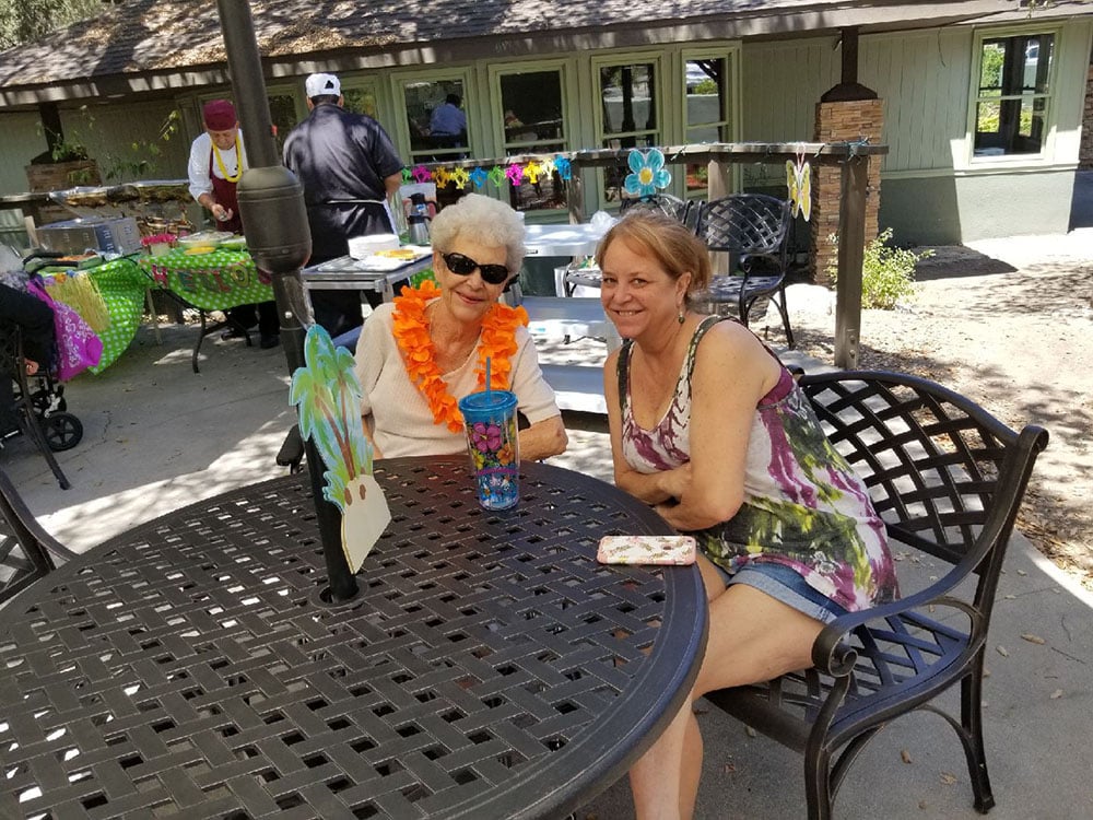 Two women sit at a metal outdoor table, smiling. One wears sunglasses and a bright orange lei; the other wears a tie-dye tank top and shorts. Colorful decorations and food tables are in the background on a sunny day.