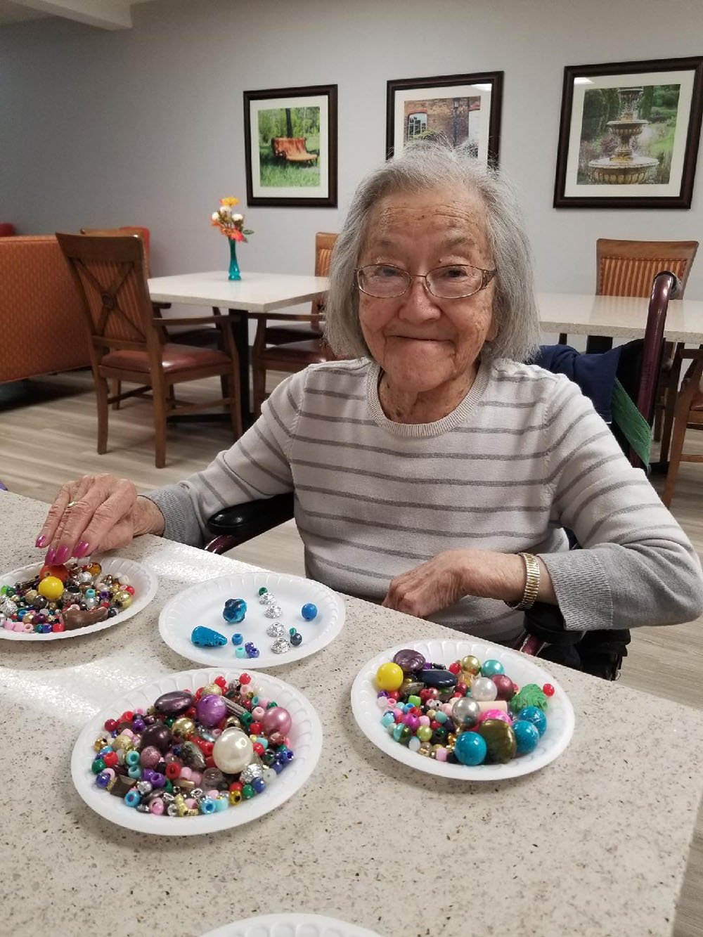 An elderly woman with glasses and gray hair smiles while sitting at a table filled with colorful beads on paper plates, in a cozy room with framed pictures and flowers in the background.