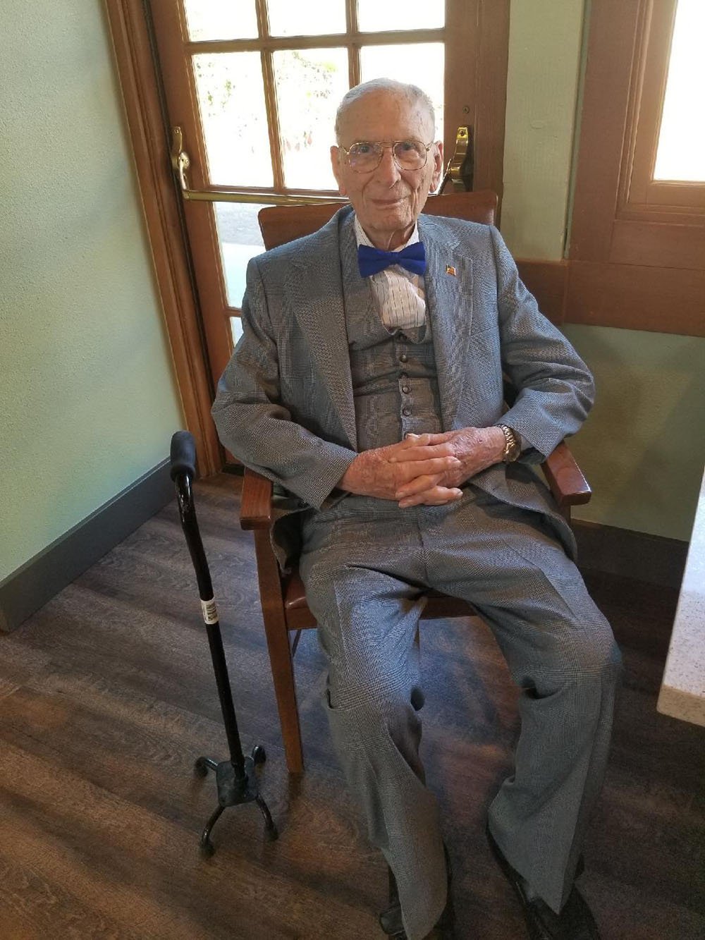 An elderly man in a gray suit and blue bow tie sits on a wooden chair indoors, hands folded in his lap. A black cane rests by his side, and sunlight streams through a window and glass door behind him.