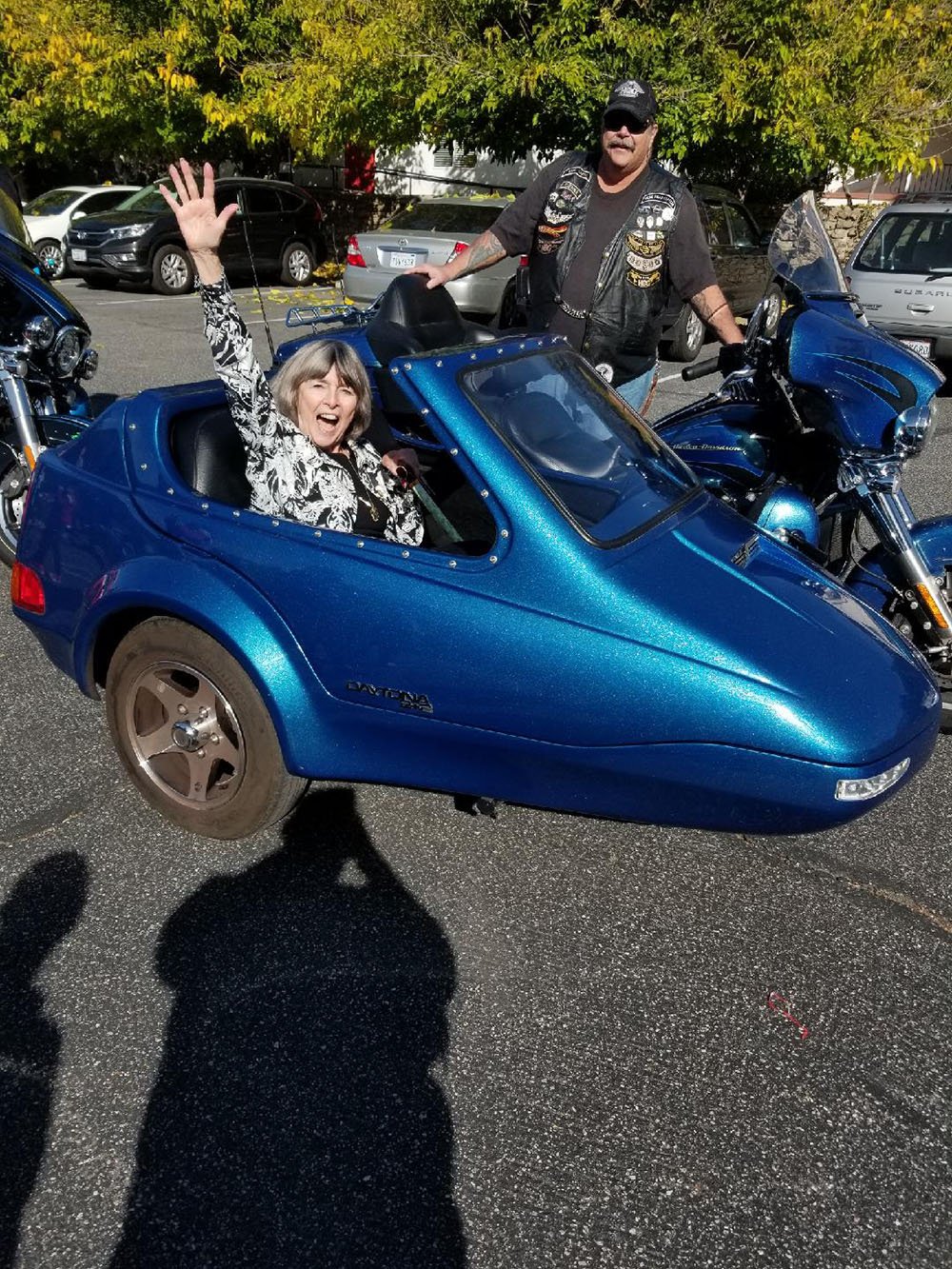 A smiling woman waves from a blue motorcycle sidecar while a man in sunglasses and a vest stands beside a motorcycle, with cars and trees in the background.
