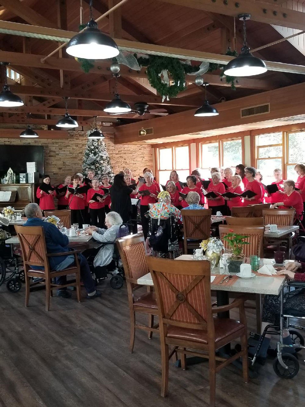 A choir dressed in red sings in front of a decorated Christmas tree to an audience of elderly people seated at tables in a cozy, wooden dining hall with large windows and festive greenery.