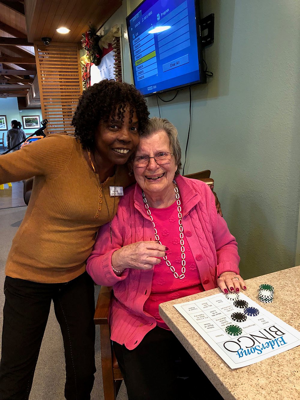 Two women smile together; one stands, the other sits at a table with a Blessing Bingo card and bingo chips. The atmosphere is cheerful, with a TV and wall art in the background.