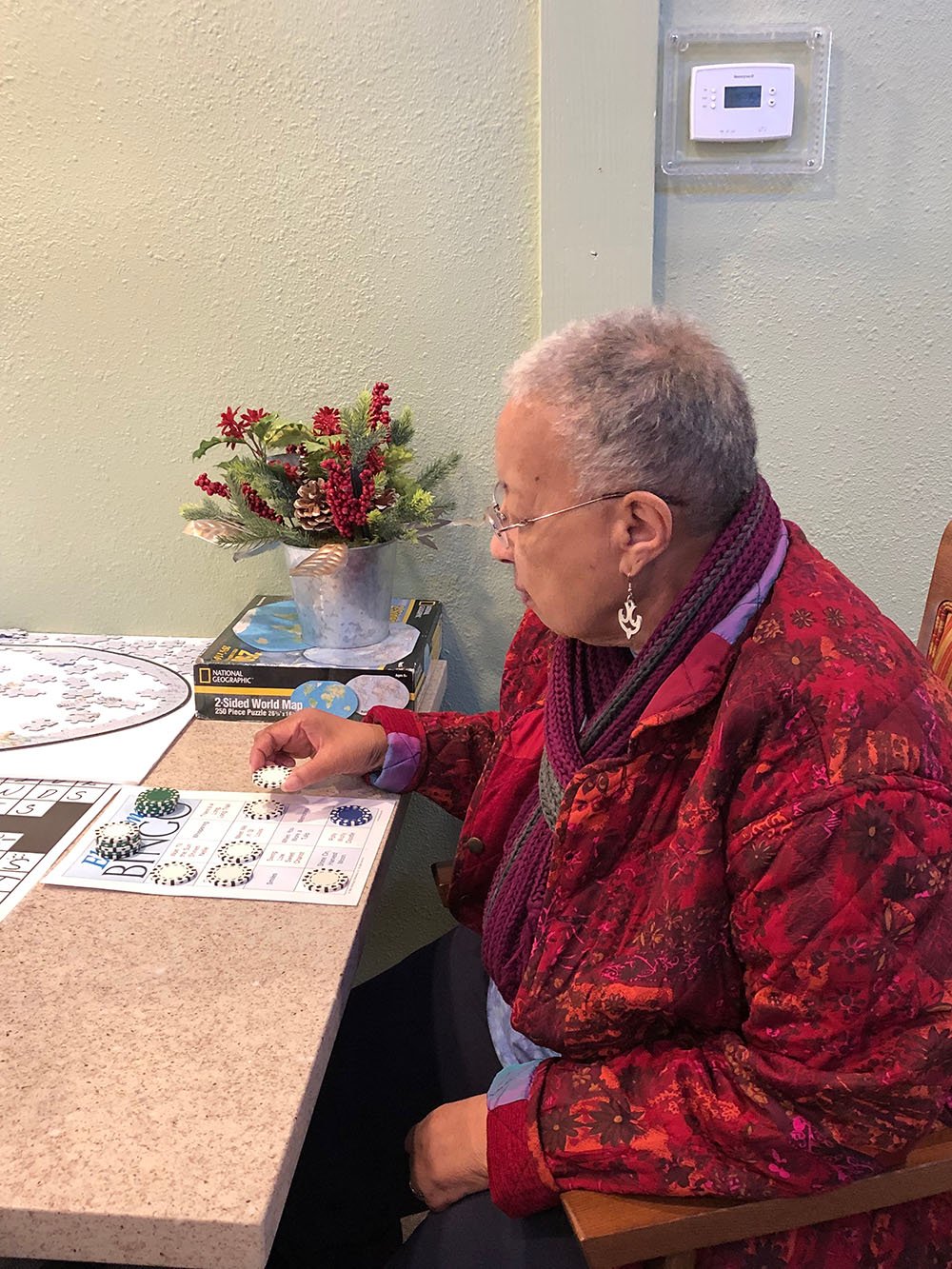 An elderly woman wearing a red patterned jacket and purple scarf sits at a table, playing a board game. A small festive plant decorates the table beside her.