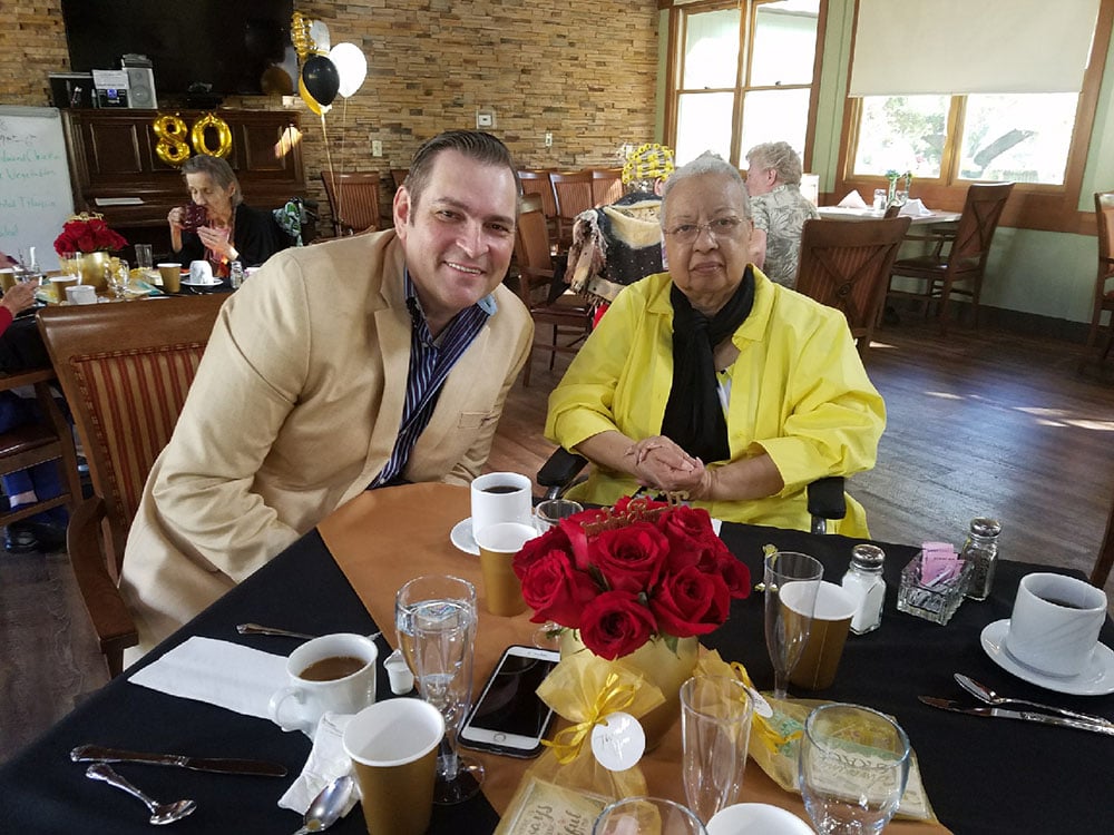 A man in a beige jacket and an older woman in a yellow outfit sit at a table with coffee cups and a bouquet of red roses, at a decorated restaurant celebrating an 80th birthday. Other guests are visible in the background.