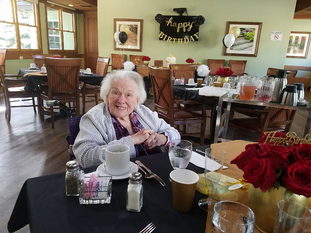 An elderly woman smiles at a table set for a celebration in a bright dining room decorated with “Happy Birthday” banners, balloons, flowers, and drinks.
