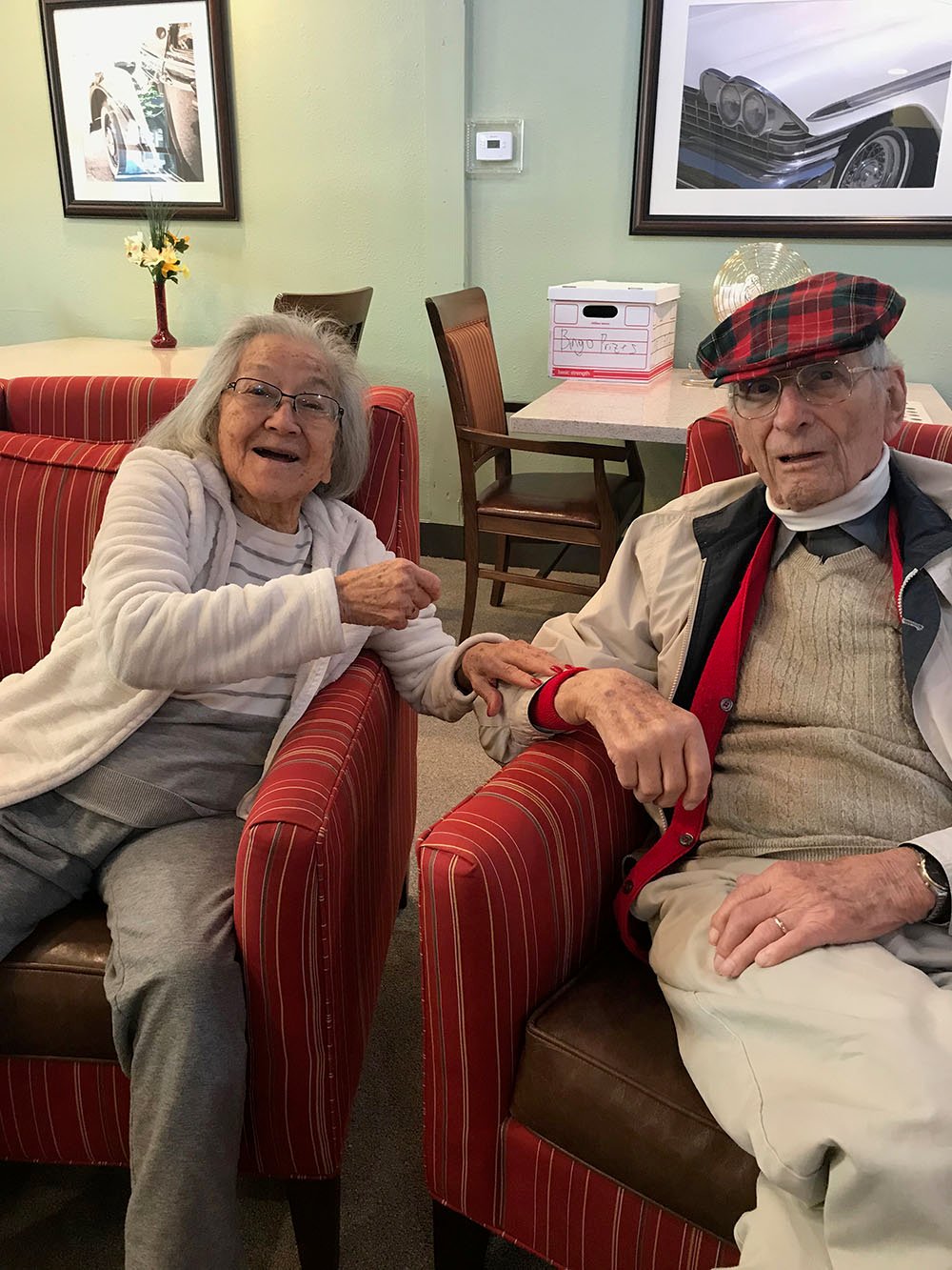 An elderly woman and man sit side by side in red-striped chairs, smiling and holding hands. The man wears a plaid cap and glasses; the woman wears glasses and a gray outfit. Framed car photos hang on the wall behind them.