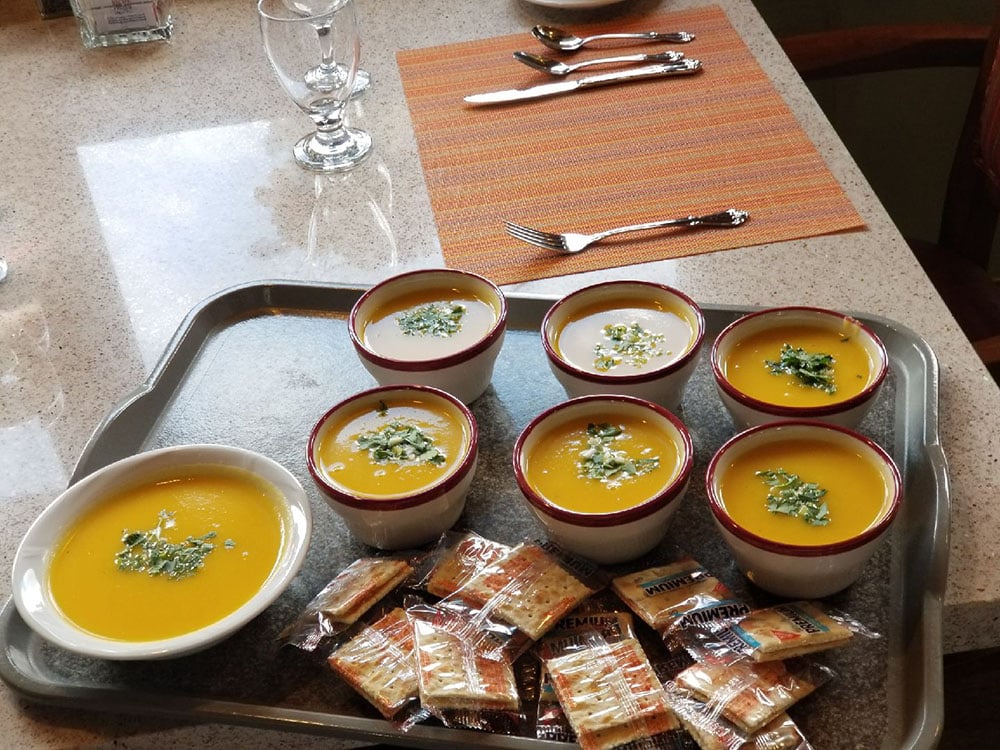 A tray with six bowls of soup, each garnished with herbs, and a bowl of crackers. The tray is set on a counter near place settings with glassware, utensils, and an orange placemat.