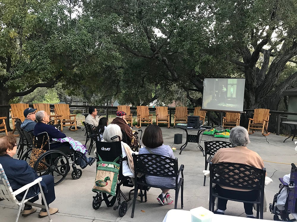 A group of older adults, some in wheelchairs, sit outside watching a movie projected onto a screen under large trees. Several empty wooden chairs are lined up in the background.