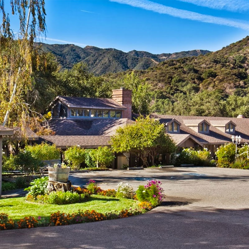 A rustic lodge with a brown roof surrounded by lush trees and colorful flowerbeds, set against a backdrop of green mountains under a blue sky with wispy clouds.