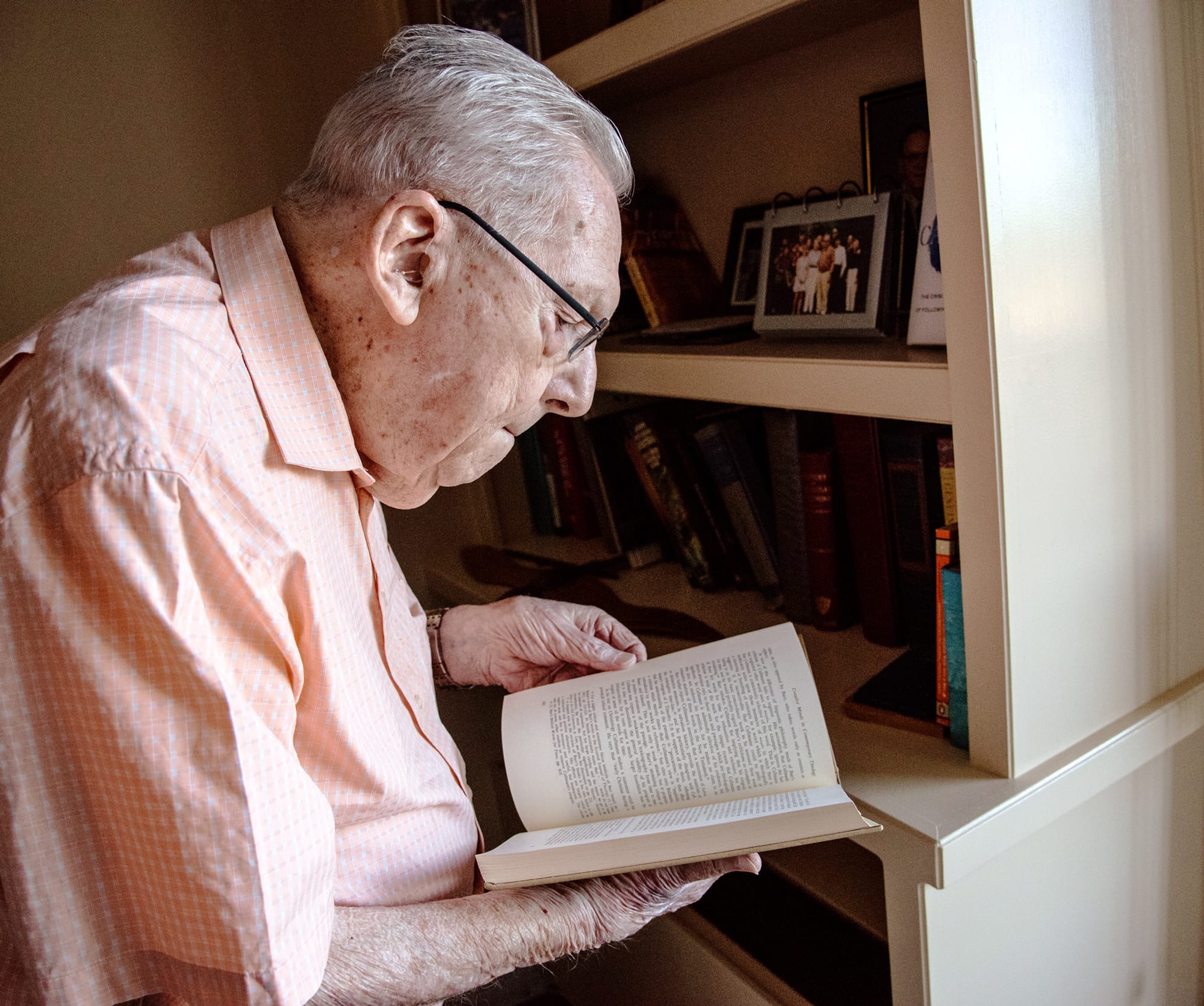 An elderly man with gray hair and glasses stands by a bookshelf, intently reading an open book. Framed photos and various books are visible on the shelves behind him.