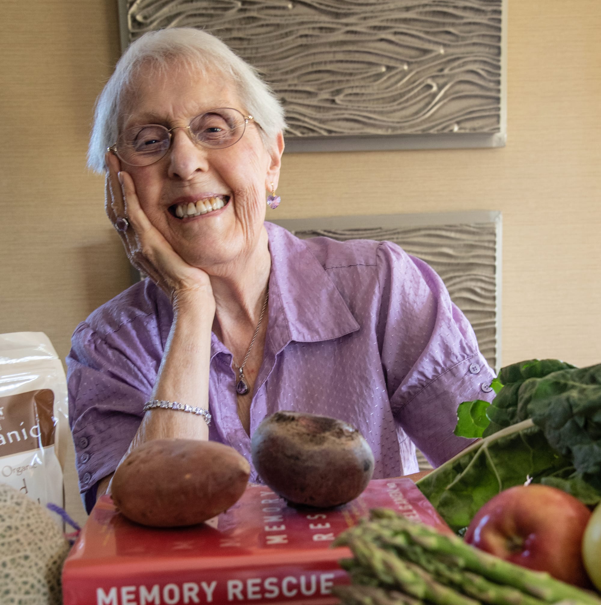 An elderly woman with short white hair and glasses smiles warmly, resting her head on her hand. She sits at a table with vegetables, fruit, and a book titled Memory Rescue in front of her.