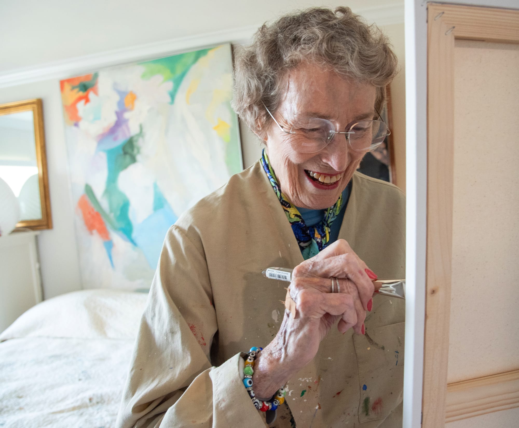 An elderly woman smiles as she paints on a canvas in a bright room, wearing glasses and a paint-stained smock. A colorful abstract painting hangs on the wall behind her.