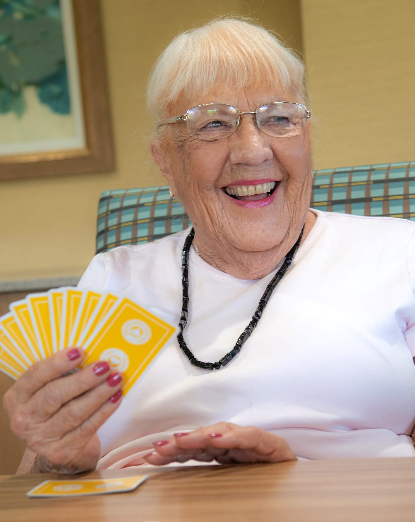 An older woman with white hair, glasses, and a black beaded necklace smiles and holds a hand of yellow playing cards while sitting at a table.