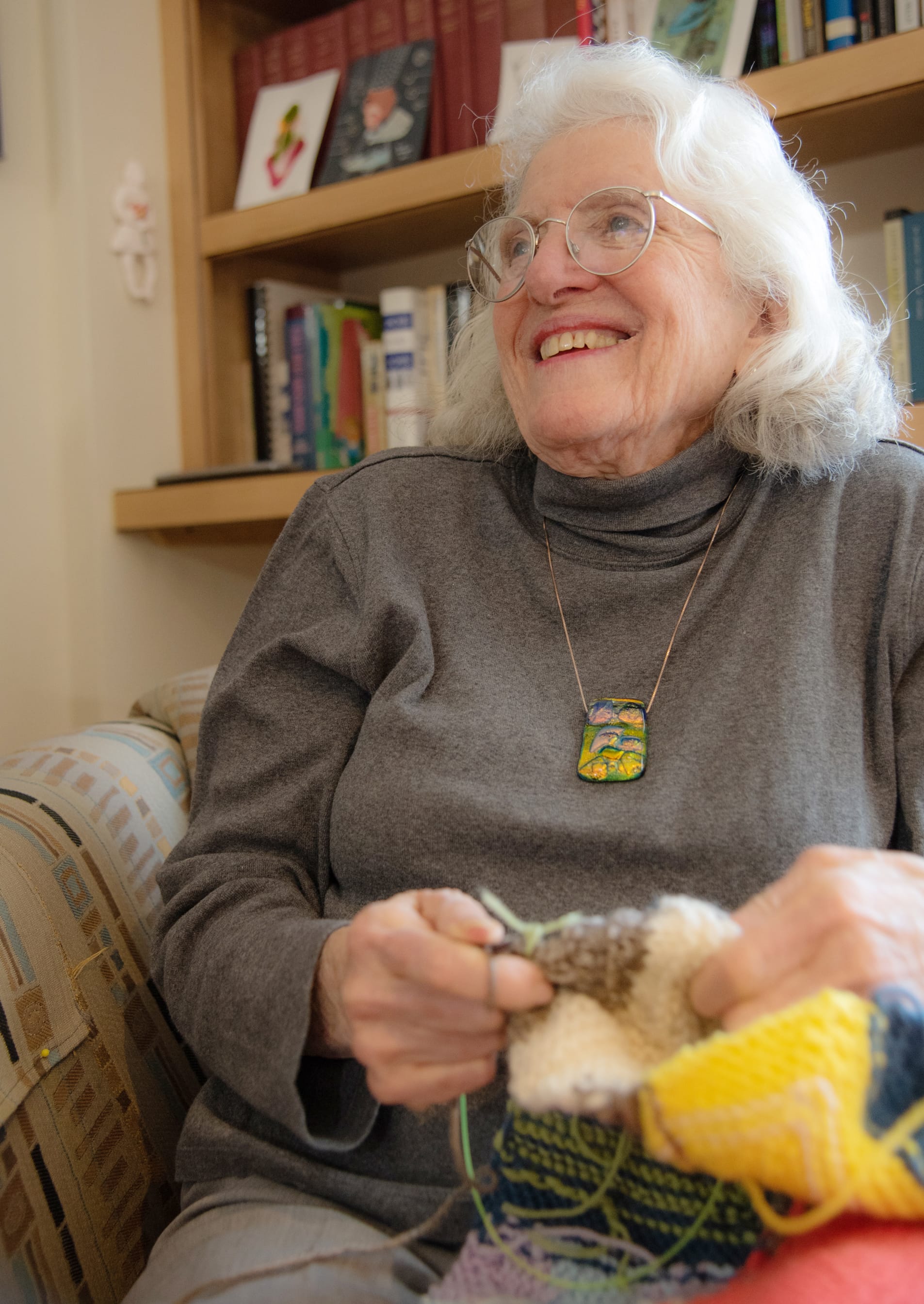 An elderly woman with white hair and glasses, wearing a gray turtleneck and colorful pendant, smiles while knitting on a patterned couch with a bookshelf in the background.