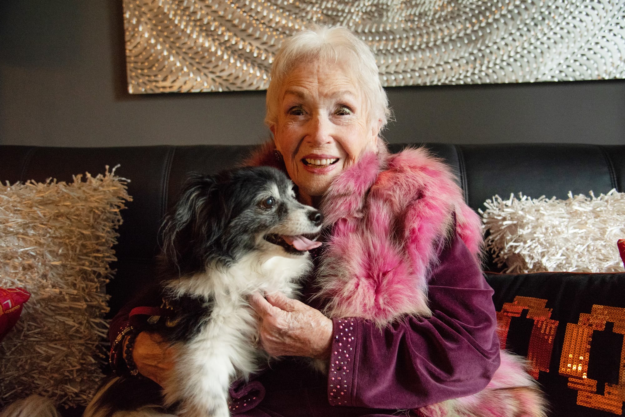 An elderly woman with short white hair, wearing a pink and purple outfit with a fur stole, smiles while sitting on a couch and holding a small black and white dog on her lap.
