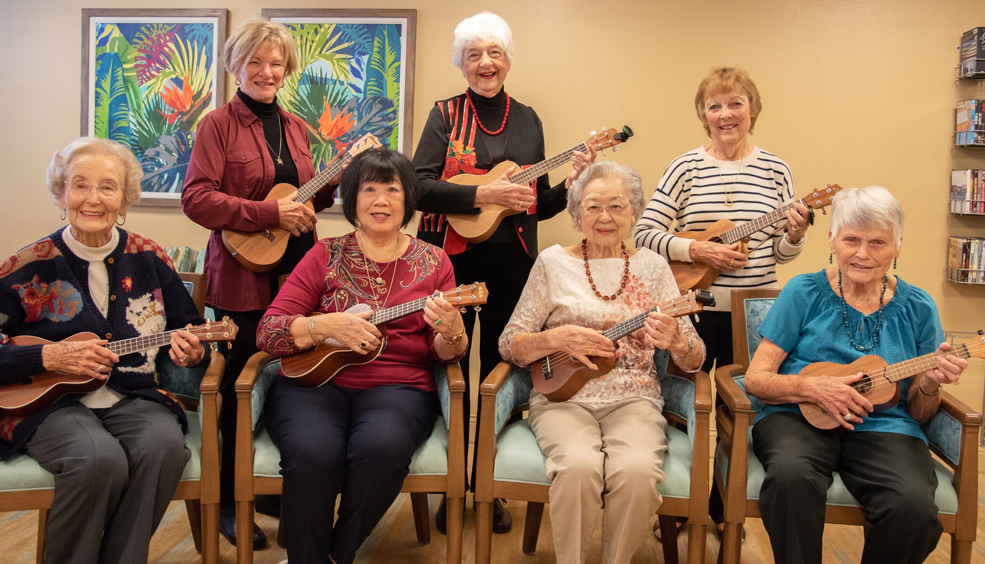Seven older women sit and stand together indoors, each holding a ukulele and smiling. The background features two colorful framed artworks and a beige wall. The atmosphere is cheerful and friendly.